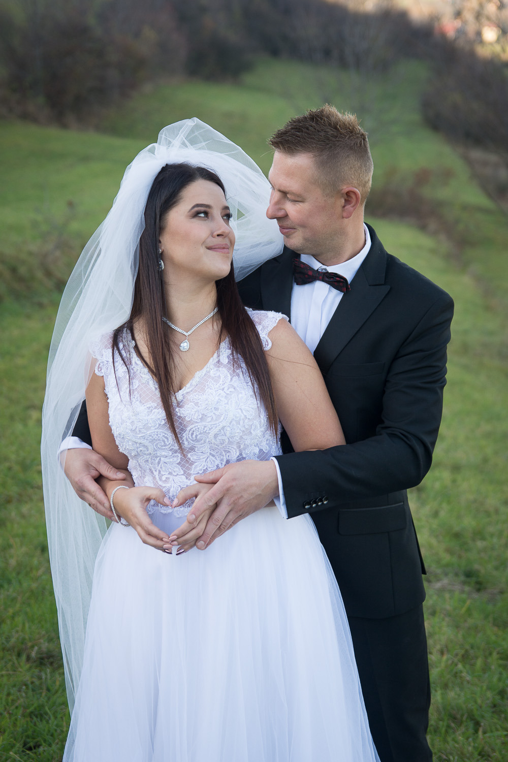 Uśmiechnięta panna młoda w białej sukni i welonie, spoglądająca na pana młodego w czarnym garniturze z muszką; oboje stoją na tle zielonego wzgórza, a ich dłonie tworzą kształt serca.