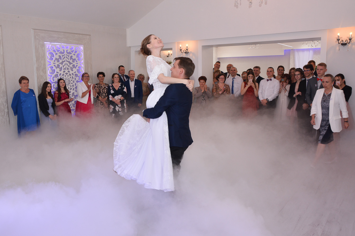 Pan młody podnosi uśmiechniętą pannę młodą na tle gości weselnych i ciężkiego dymu na parkiecie.