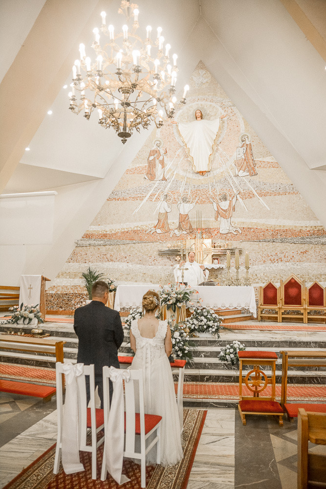 Para młoda stoi przed ołtarzem w kościele podczas ceremonii ślubnej, widoczny ksiądz, dekoracje kwiatowe i mozaika religijna na ścianie