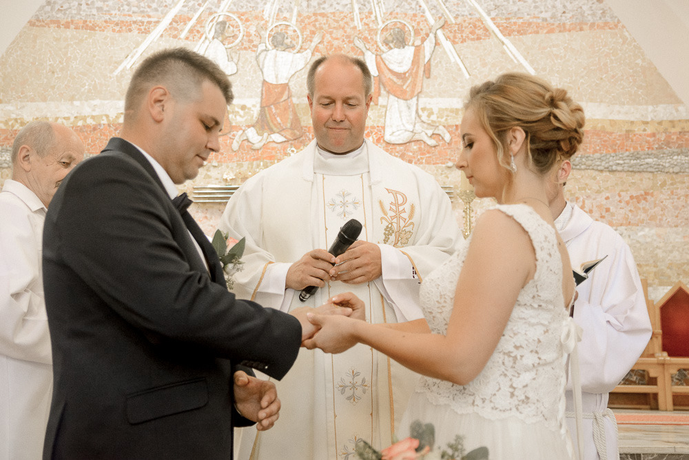 Para młoda wymienia obrączki podczas ceremonii ślubnej w kościele, w tle mozaika z motywem religijnym, ksiądz z mikrofonem.