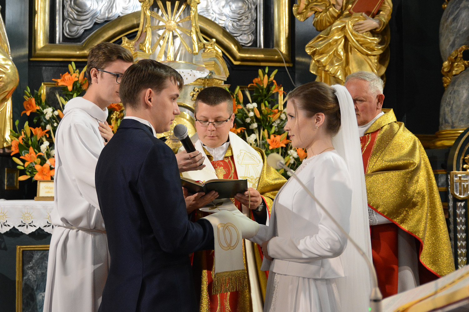 Para młoda podczas ceremonii ślubnej w kościele, z księdzem odczytującym przysięgę małżeńską, na tle bogato zdobionego ołtarza i kwiatów.