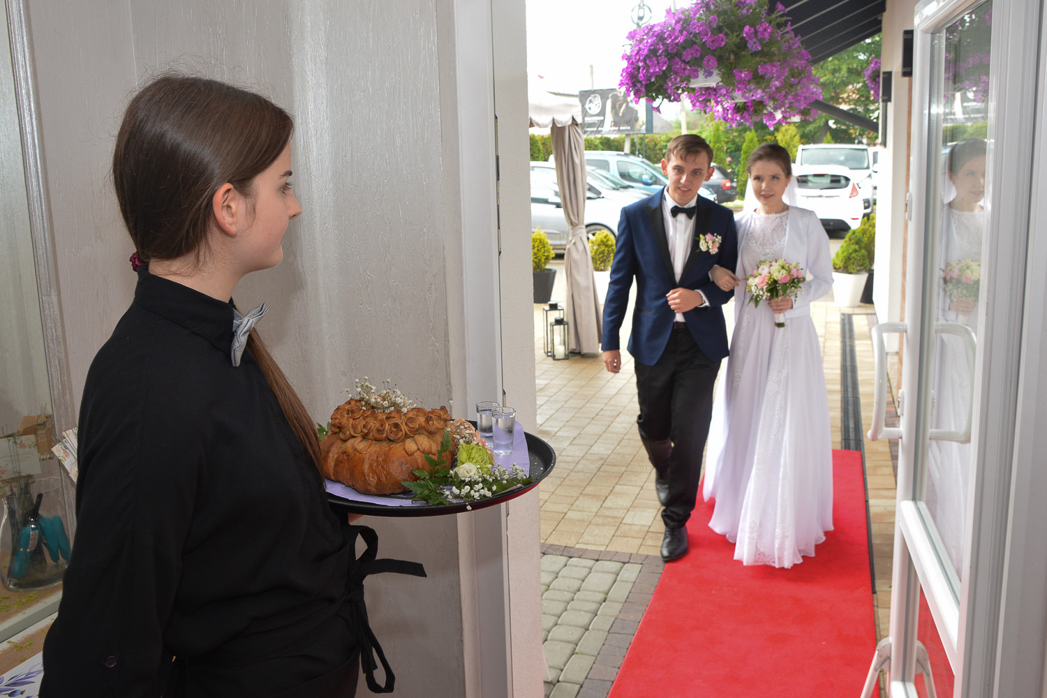 Panna młoda i pan młody wchodzą do sali weselnej, witani przez osobę z chlebem weselnym na tacy.