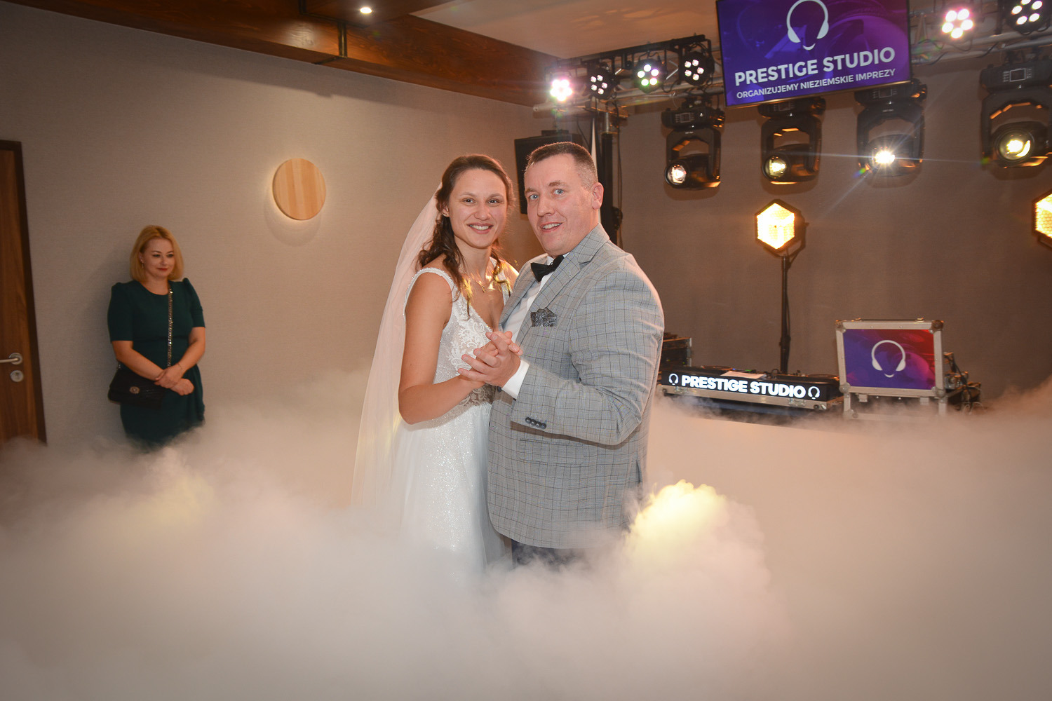 Para młoda w tańcu w chmurze dymu na tle sprzętu DJ-skiego Prestige Studio, z gościem w tle po lewej stronie.