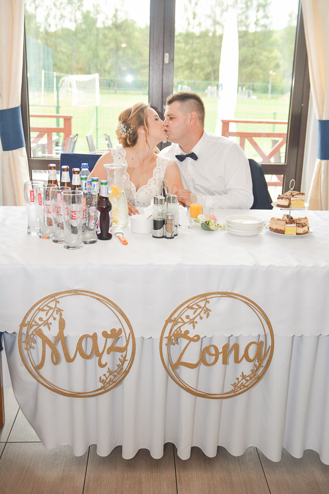Nowożeńcy całują się za stołem podczas wesela, na stole napoje, ciasta i dekoracje 'Mąż' i 'Żona'.