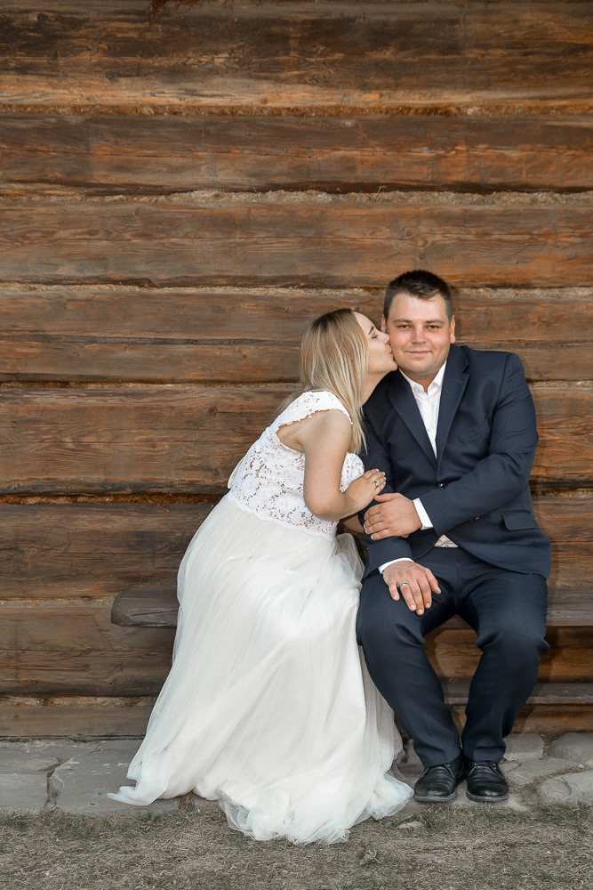 Panna młoda w koronkowej sukni całuje pana młodego w ciemnym garniturze na tle drewnianej ściany.