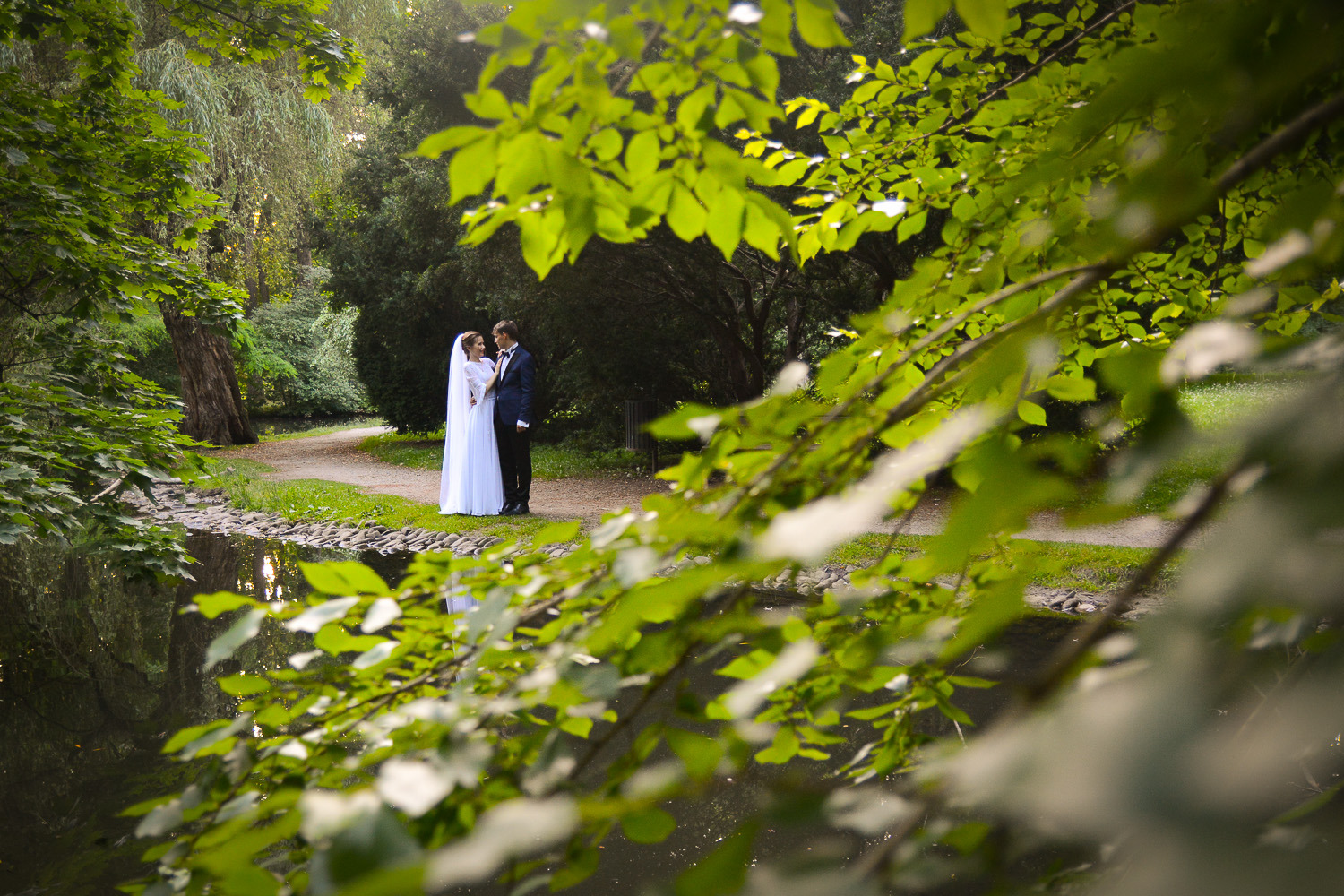 Para młoda w parku, odbicie w wodzie, gałęzie z liśćmi w pierwszym planie, on w granatowym garniturze, ona w białej sukni ślubnej i welonie.
