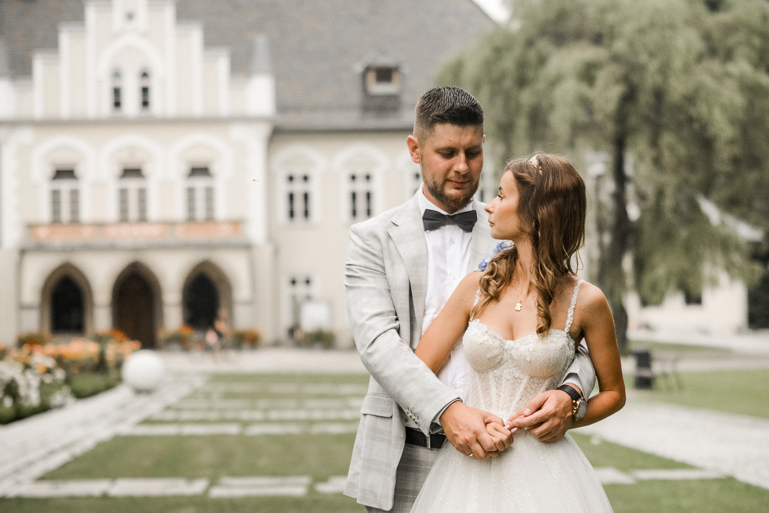 Para młoda w jasnych strojach, trzymająca się za ręce na tle zabytkowego pałacu z arkadami i zadbanego ogrodu.