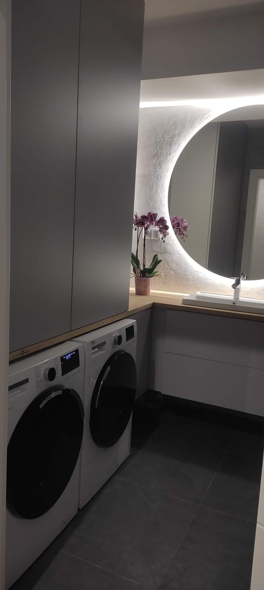 Nowoczesna łazienka z dwoma pralkami, szarą szafą, okrągłym lustrem z podświetleniem LED i fioletowym storczykiem.