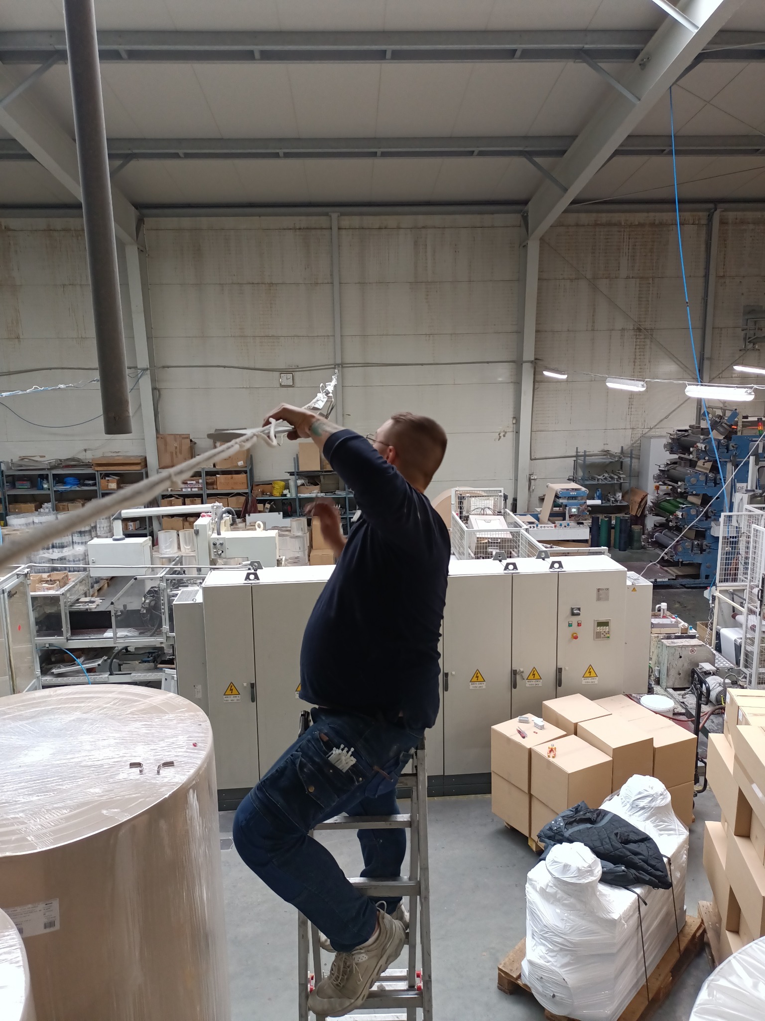 Mężczyzna na drabinie mocuje przewody w hali produkcyjnej z maszynami i materiałami.