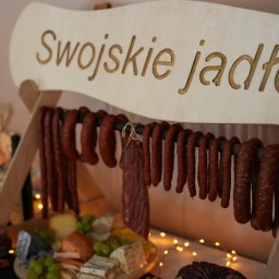 Smacznego-Domowego Jarosław Smażyk - Stół z drewnianym napisem 'Swojskie jadło', wędlinami, serami i owocami. Dekoracja świetlna w tle. Ujęcie z bliska, ciepłe światło.