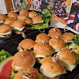 Smacznego-Domowego Jarosław Smażyk - Mini burgery z serem i warzywami na czarnych tacach, dekorowane motywem Avengers, idealne na przyjęcie tematyczne. Sezam na bułkach, świeża sałata i arbuz.