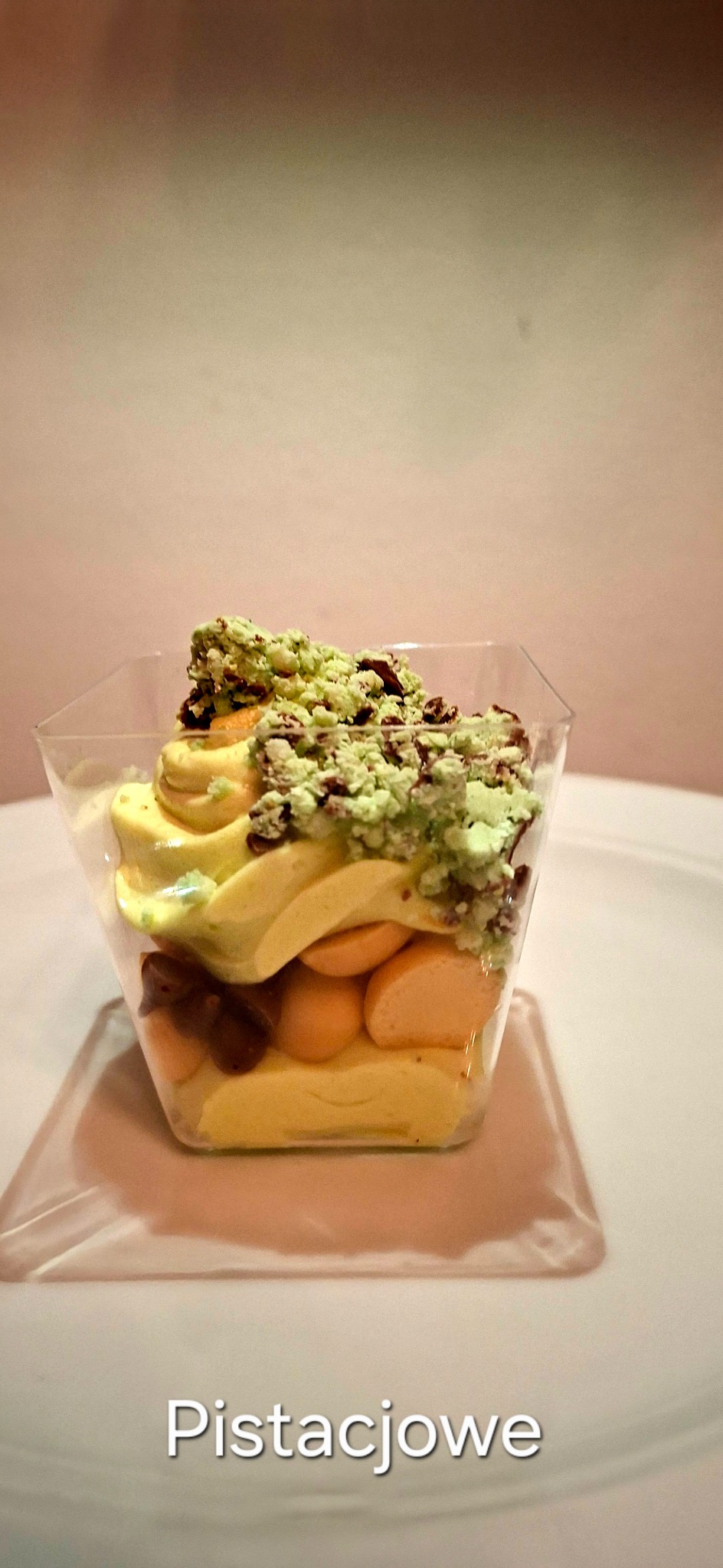 Deser pistacjowy w przezroczystym kubeczku: warstwy kremu, biszkoptów i posypki pistacjowej. Widoczne detale tekstury i koloru, na białym talerzu, delikatne oświetlenie.