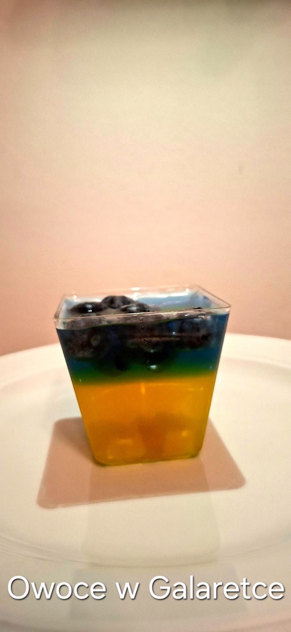 Deser w przezroczystym kubeczku: żółta galaretka z owocami, zielona warstwa i borówki w niebieskiej galaretce. Całość na białym talerzu.