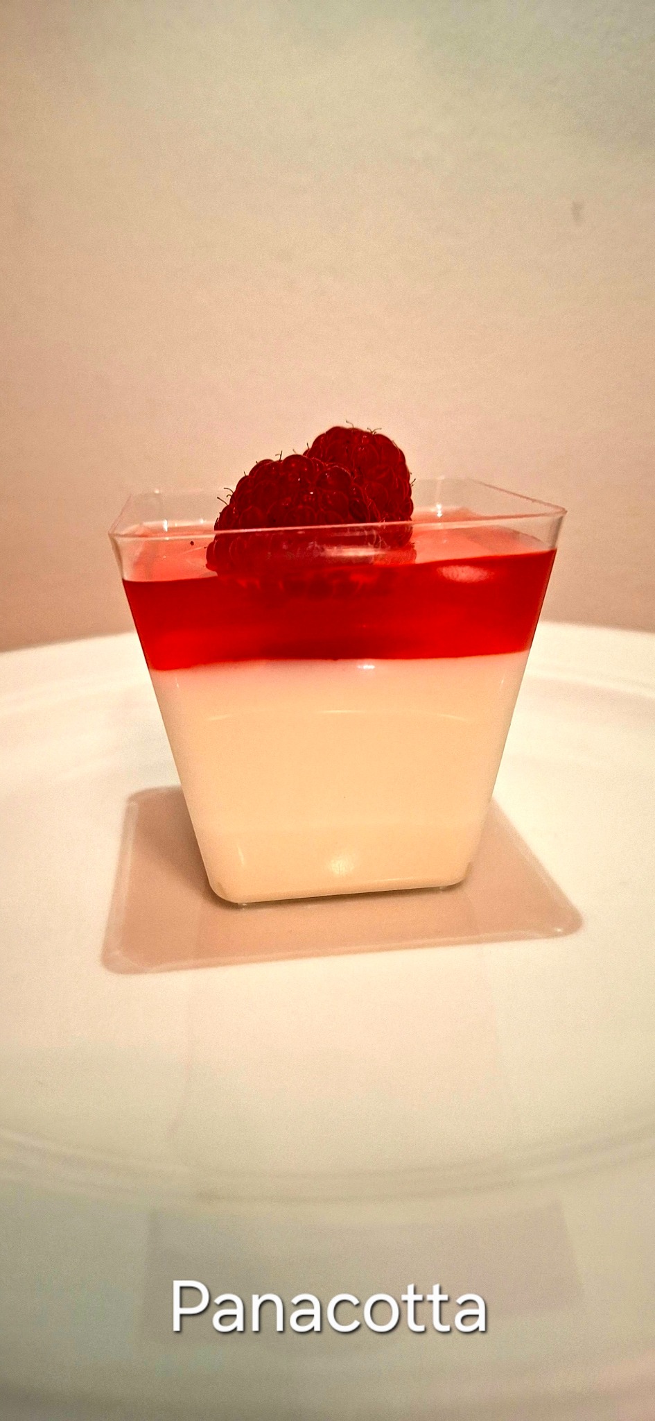 Panna cotta w przezroczystym, kwadratowym naczyniu, udekorowana czerwoną galaretką i malinami, na białym talerzu. Delikatne oświetlenie podkreśla kremową konsystencję.