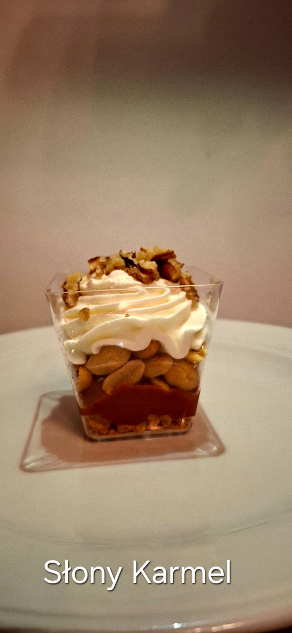 Deser 'Słony Karmel' w przezroczystym kubeczku: warstwy karmelu, orzechów, kremu. Całość na białym talerzu, delikatne oświetlenie.