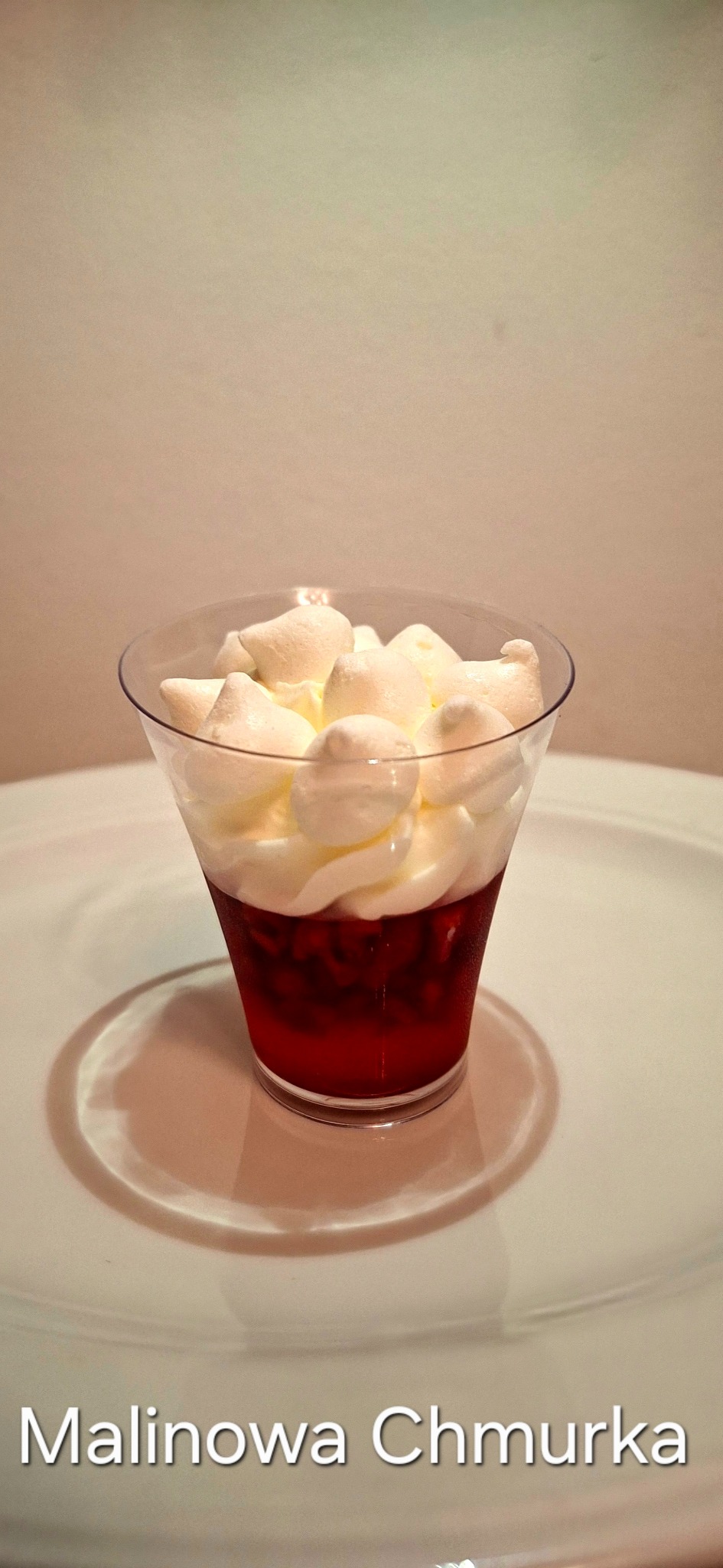 Deser malinowy w przezroczystym kubeczku na białym talerzu, zwieńczony bitą śmietaną i bezami. Widoczne delikatne cienie i kremowe tło.