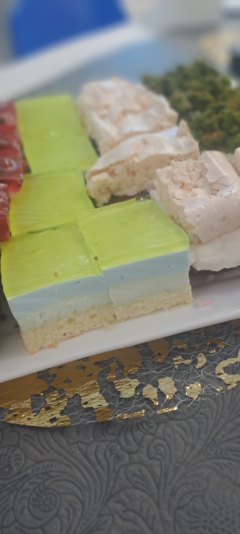 Kawałki ciasta z galaretką, bezą i zielonymi posypkami na białym talerzu z dekoracyjnym obrusem. Widoczne warstwy i tekstury ciasta.