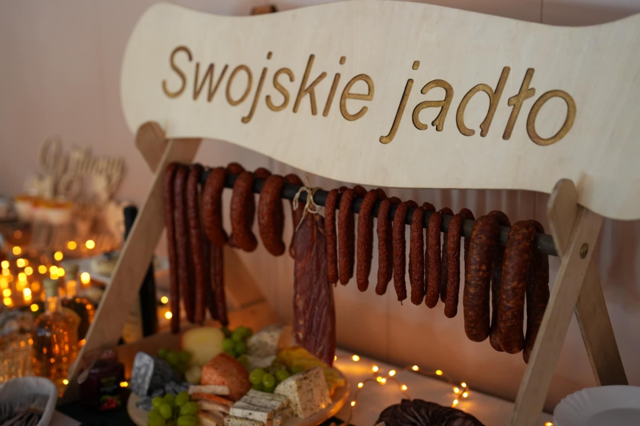 Stół z drewnianym napisem 'Swojskie jadło', wędlinami, serami i owocami. Dekoracja świetlna w tle. Ujęcie z bliska, ciepłe światło.