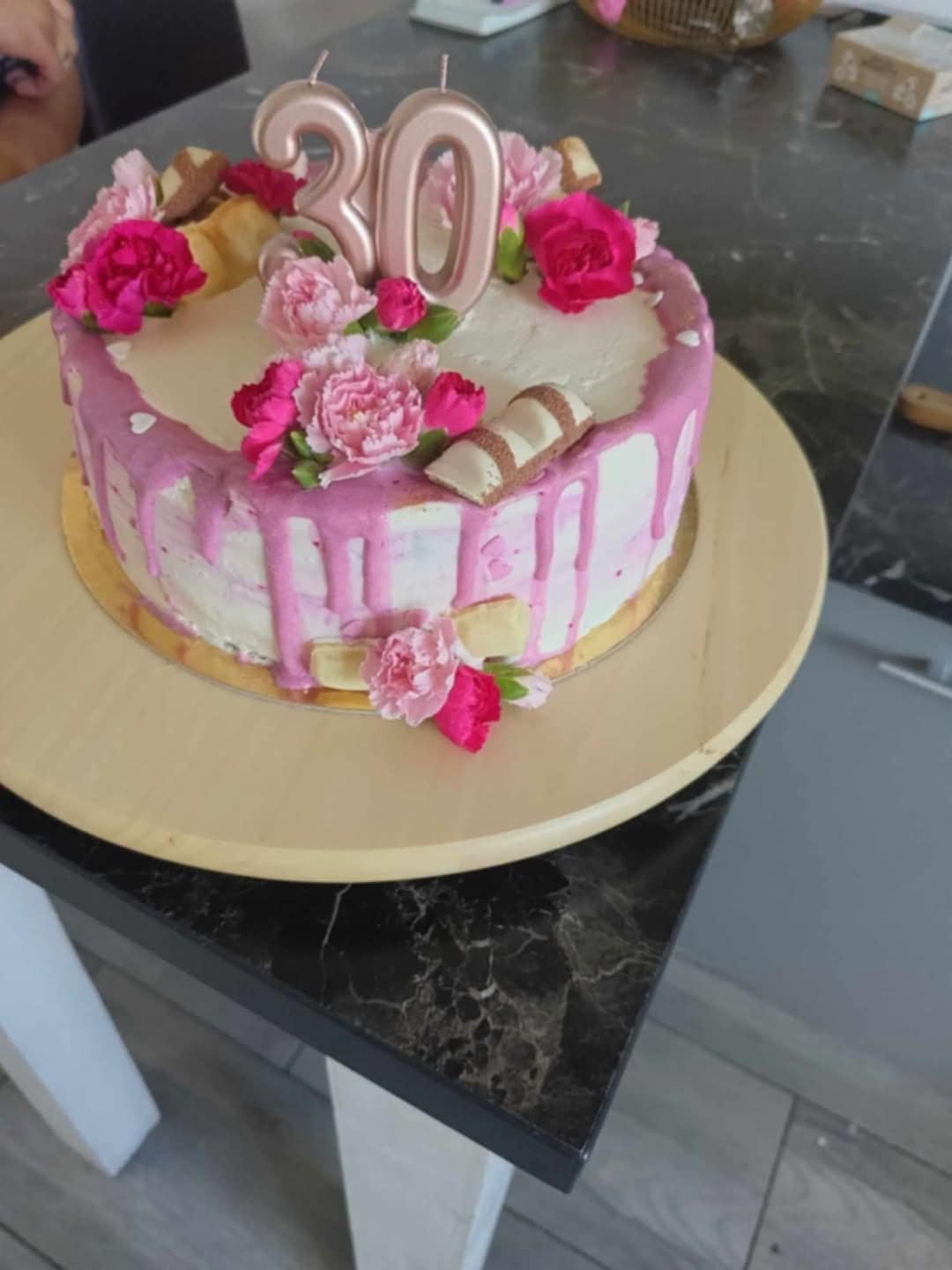 Tort urodzinowy z różowym lukrem, kwiatami i świeczkami w kształcie liczby 30, umieszczony na drewnianym talerzu na tle marmurowego blatu.