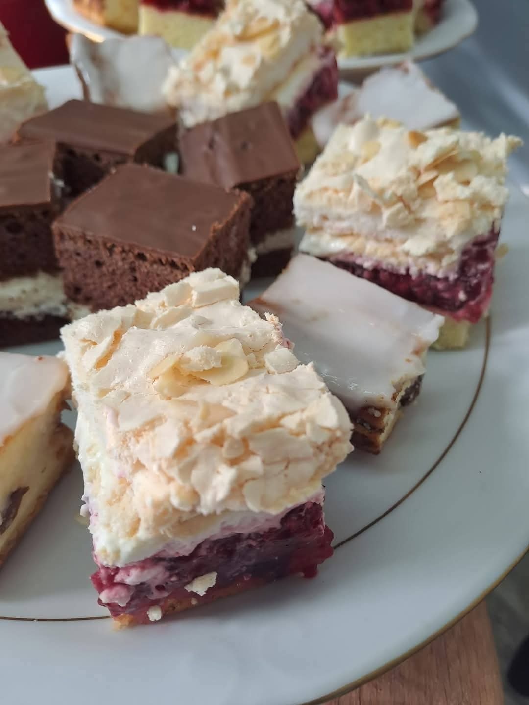 Różnorodne ciasta na białym talerzu z delikatnym złotym obramowaniem. Wśród nich wyróżniają się ciasta z kremem, owocami i czekoladową polewą.