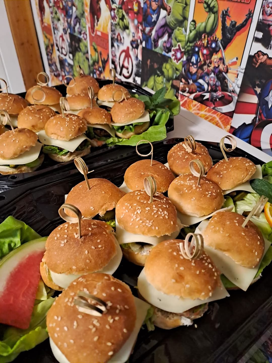 Mini burgery z serem i warzywami na czarnych tacach, dekorowane motywem Avengers, idealne na przyjęcie tematyczne. Sezam na bułkach, świeża sałata i arbuz.
