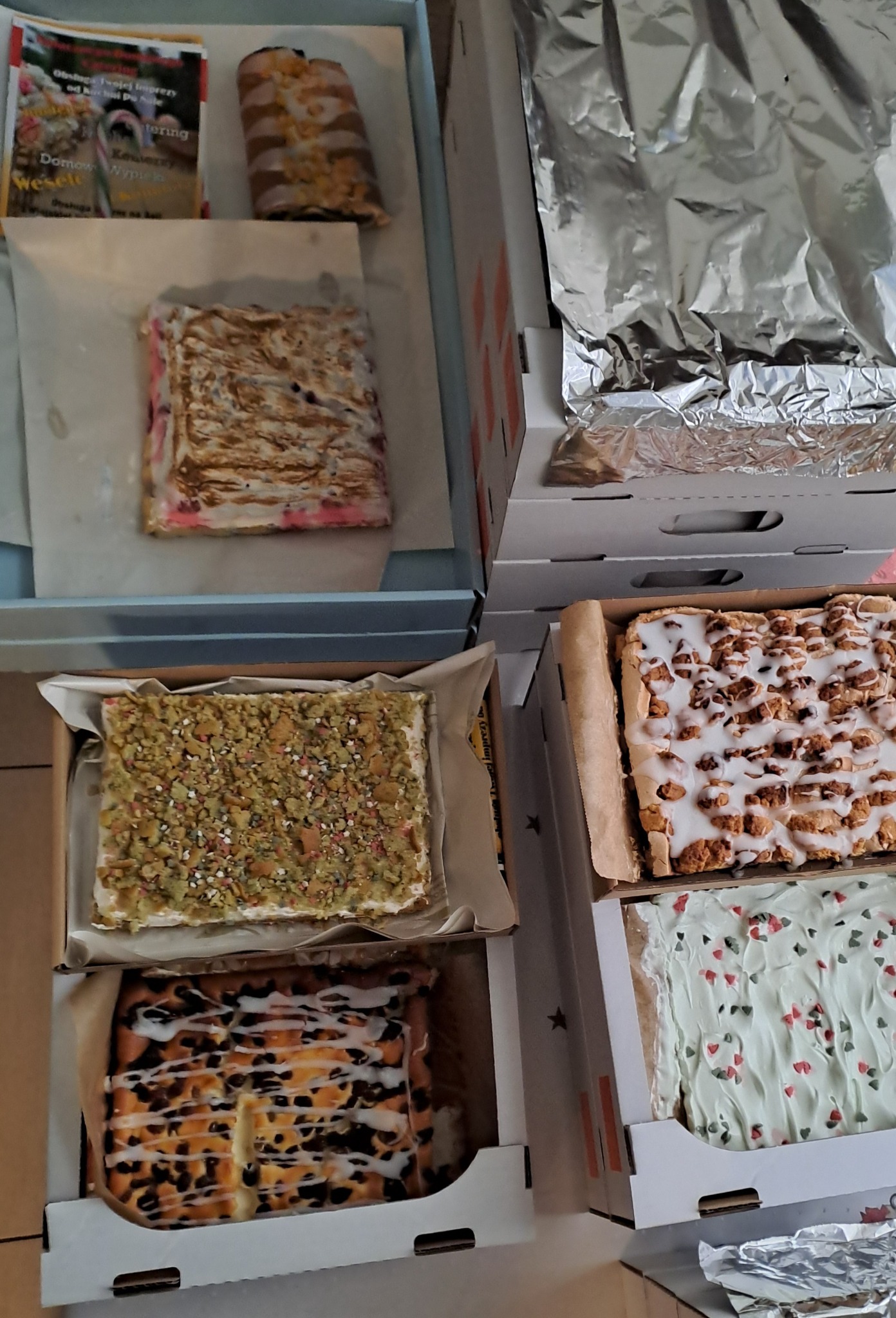 Różnorodne ciasta w kartonowych pudełkach, przygotowane w Bydgoszczy, gotowe do transportu, widoczne różne polewy i posypki, w tym pistacjowa, czekoladowa i owocowa.