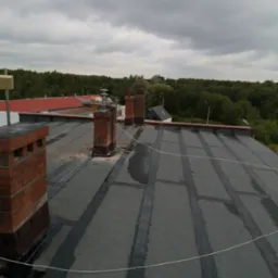Widok z góry na płaski dach pokryty papą z widocznymi kominami z cegły i sznurkiem zabezpieczającym, w tle zalesiony teren i pochmurne niebo.