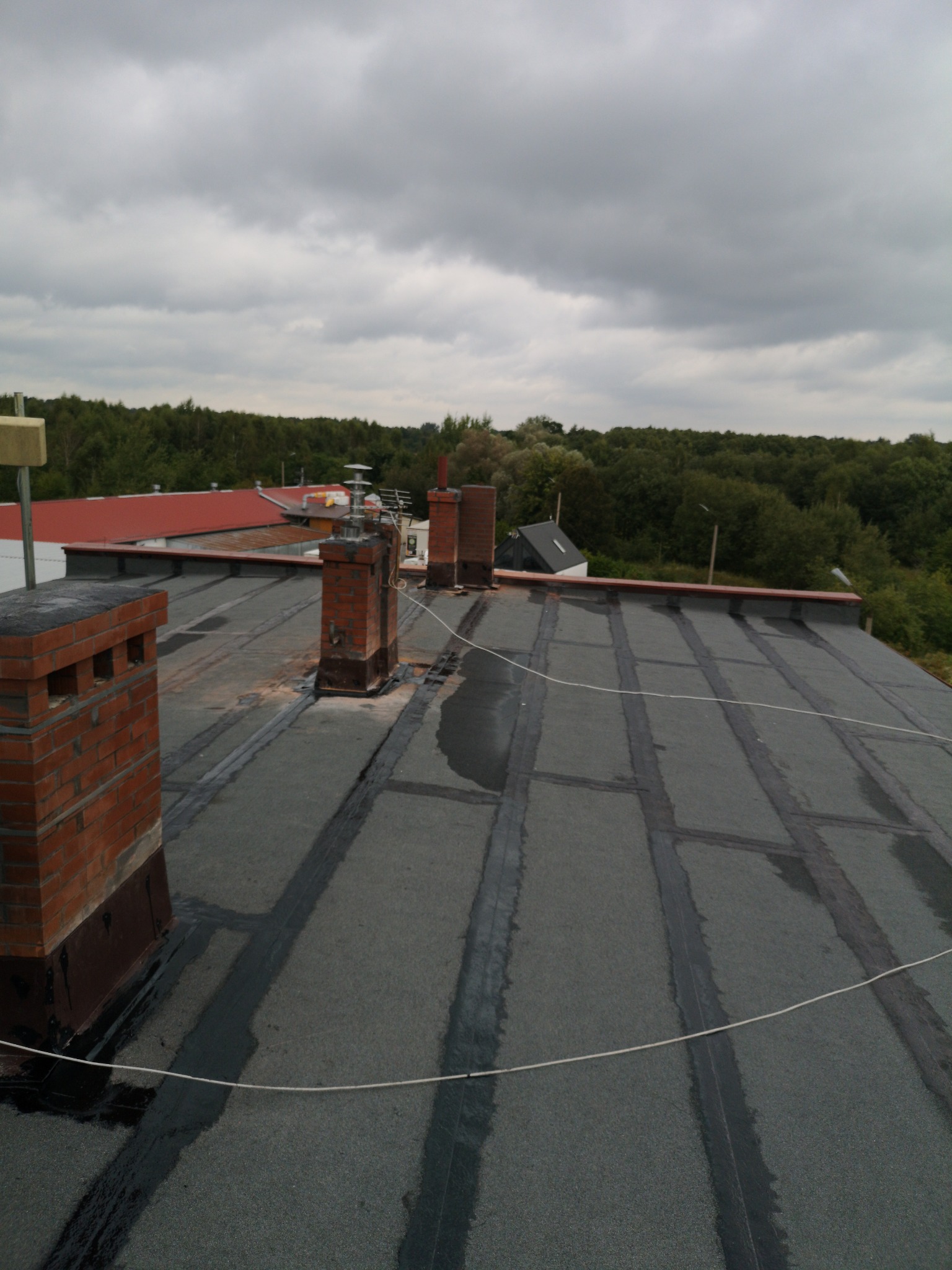 Widok z góry na płaski dach pokryty papą z widocznymi kominami z cegły i sznurkiem zabezpieczającym, w tle zalesiony teren i pochmurne niebo.
