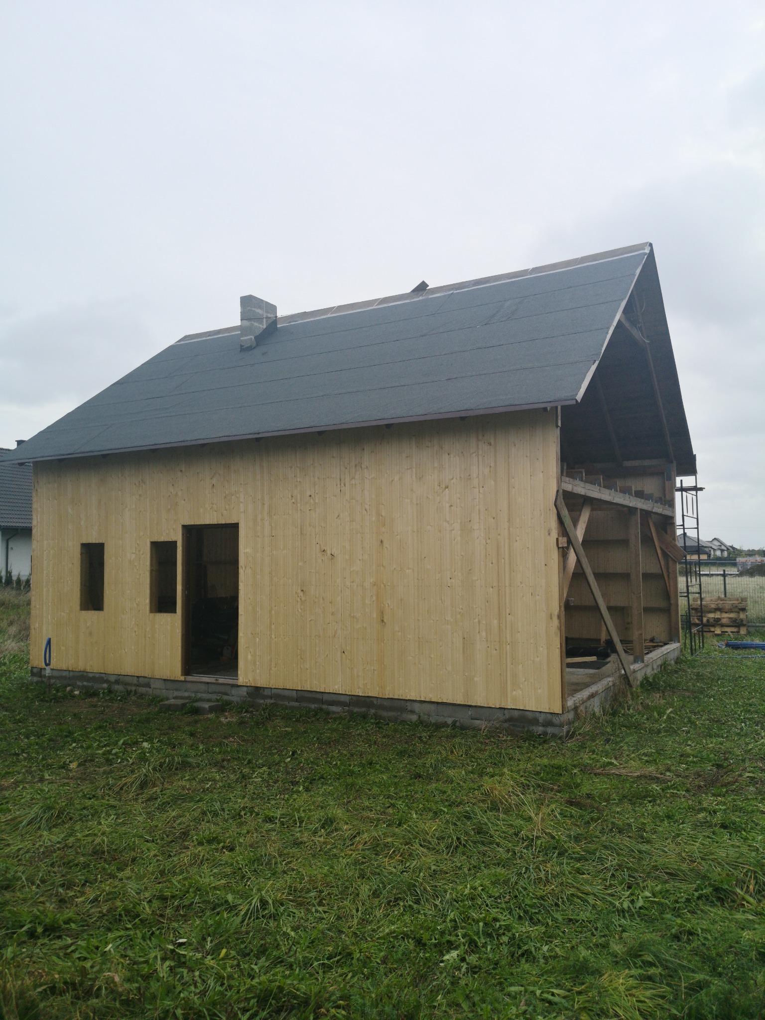 Budowa domu w trakcie: widoczna drewniana konstrukcja ścian z wyciętymi otworami okiennymi i drzwiowymi, dach pokryty papą, komin murowany, na zewnątrz trawa i pochmurne niebo.