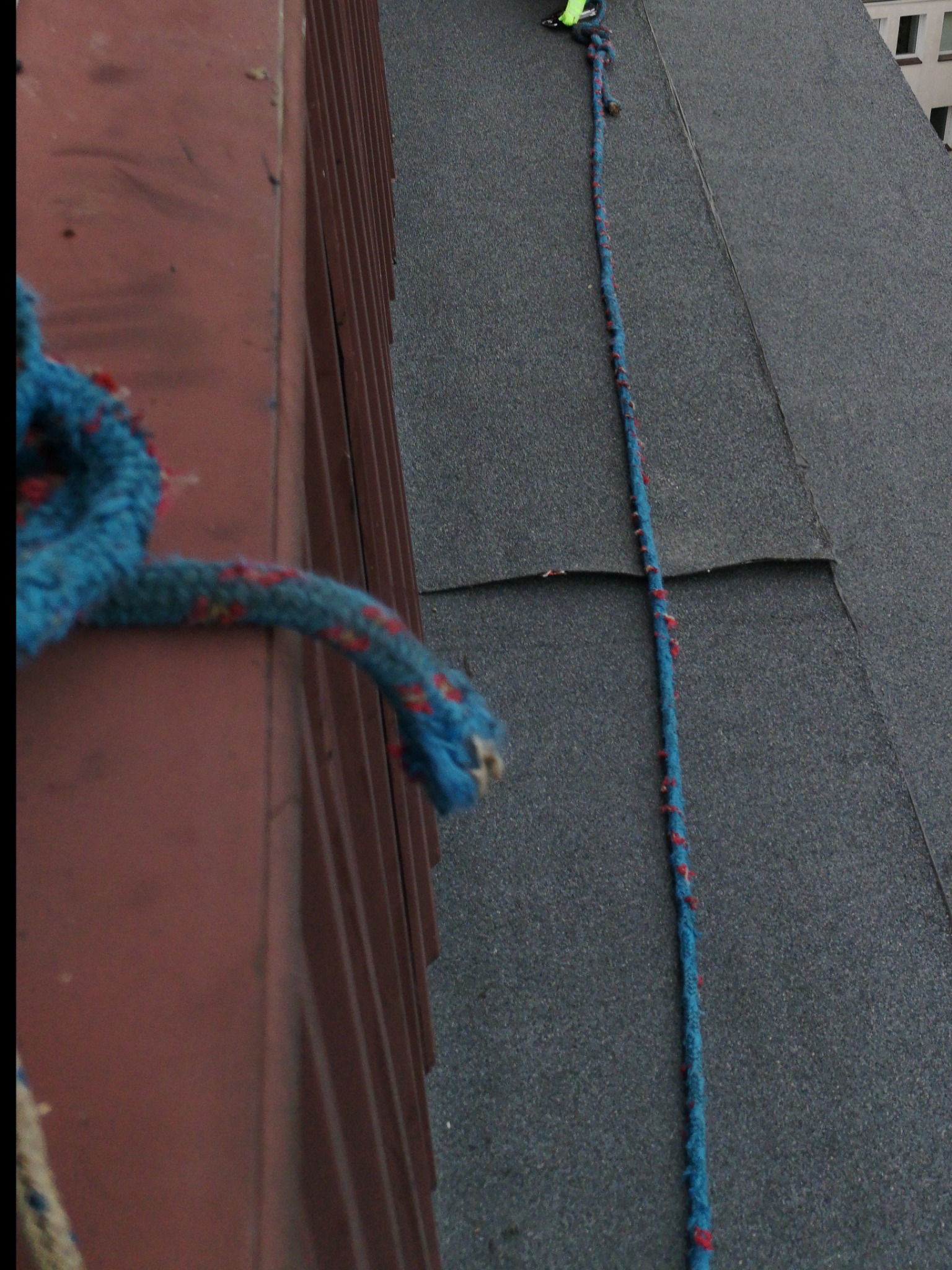 Fragment dachu pokrytego papą z widoczną liną zabezpieczającą w kolorze niebieskim z czerwonymi akcentami, leżącą na powierzchni, oraz fragmentem metalowej obróbki blacharskiej w kolorze ceglastym.