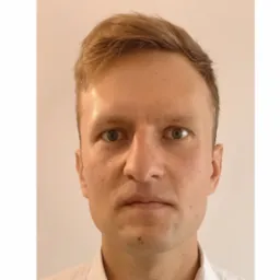 Portret mężczyzny z krótkimi blond włosami, patrzącego prosto w obiektyw, na jasnym tle.