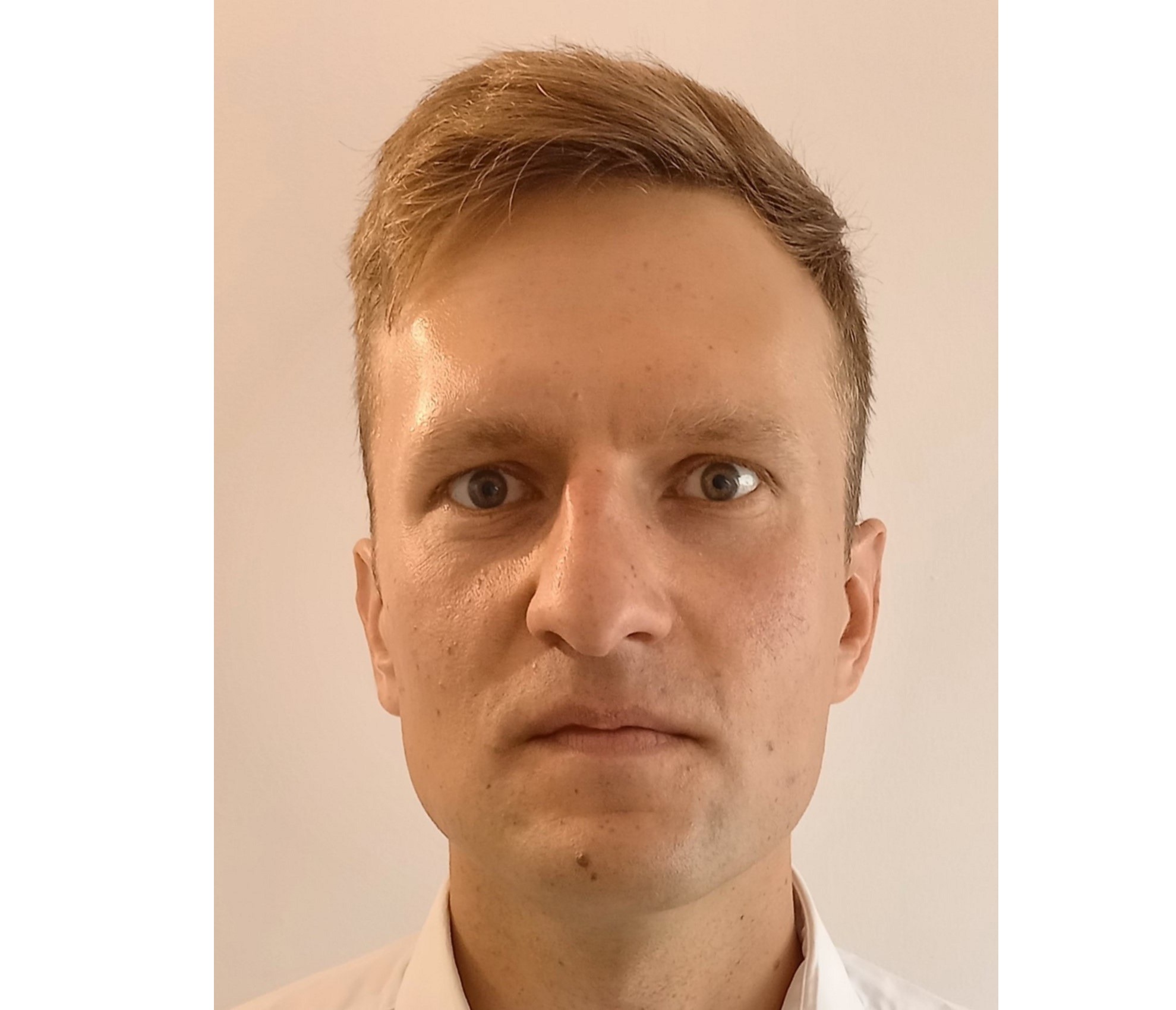 Portret mężczyzny z krótkimi blond włosami, patrzącego prosto w obiektyw, na jasnym tle.