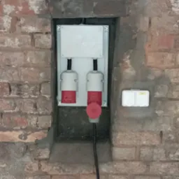 Gniazda siłowe (czerwone wtyczki) zamontowane w puszce na ceglanej ścianie z widocznymi śladami zaprawy, obok gniazdko podwójne.