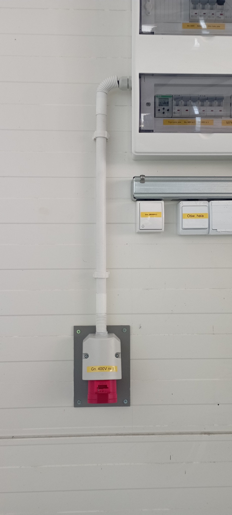 Gniazdo siłowe 400V z czerwonym wtykiem, zamontowane na szarej płycie na białej ścianie z poziomych paneli, obok tablica rozdzielcza z bezpiecznikami i przyciskami oświetlenia.