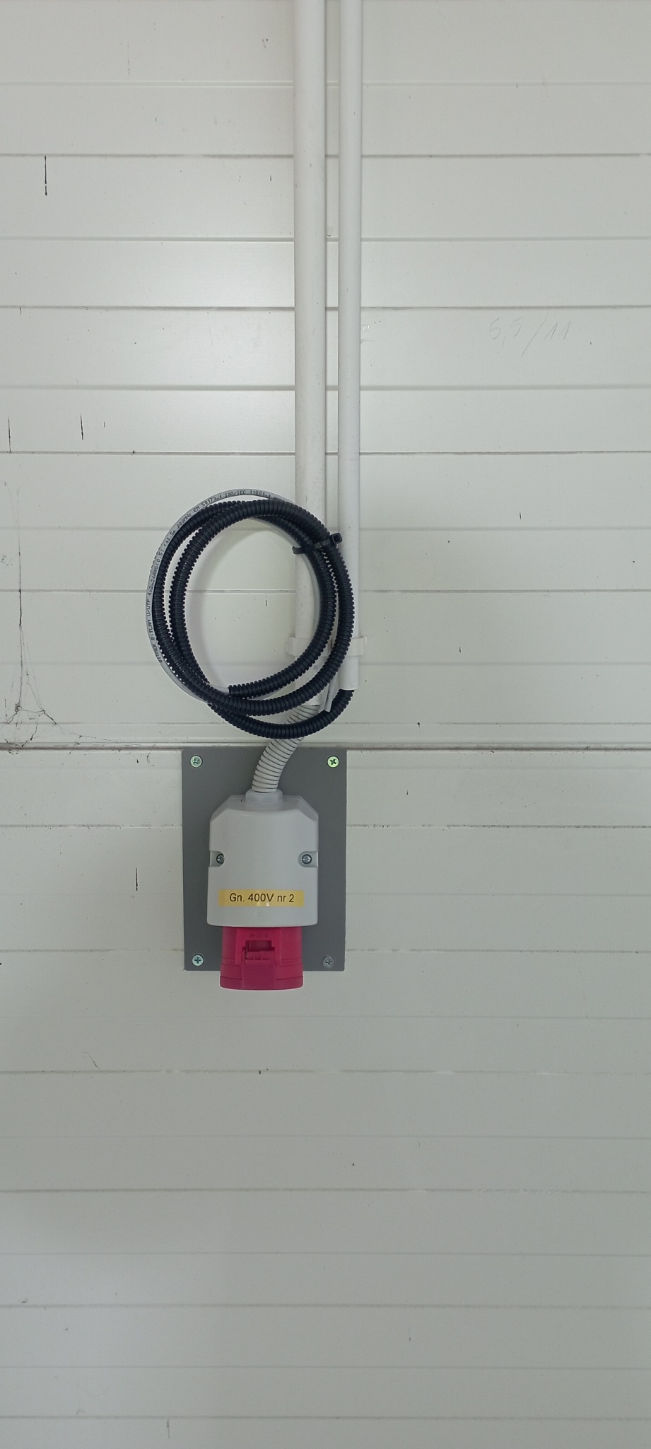 Zamontowane gniazdo siłowe 400V z podłączonym przewodem w peszlu, widoczne rury instalacyjne na tle białej ściany z paneli.