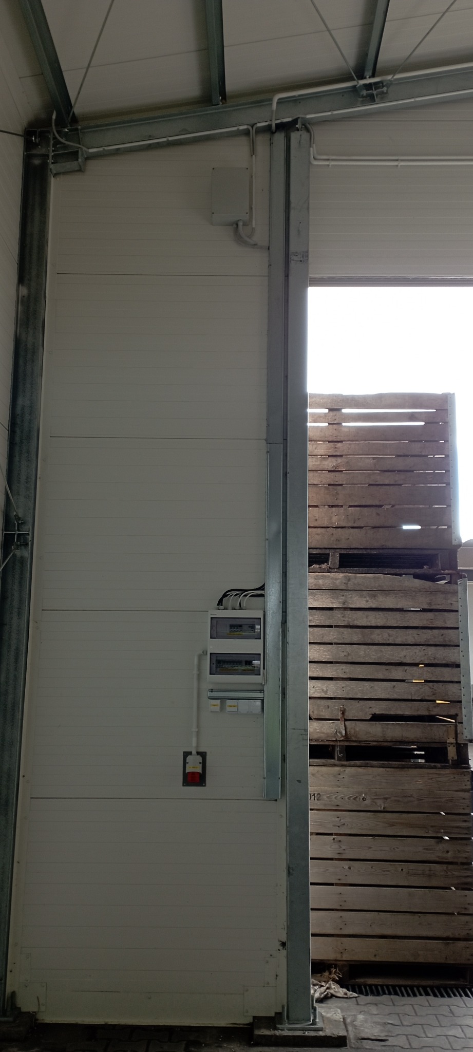 Rozdzielnica elektryczna z gniazdem siłowym zamontowana na panelowej ścianie hali magazynowej, widoczne kable i rury instalacyjne.