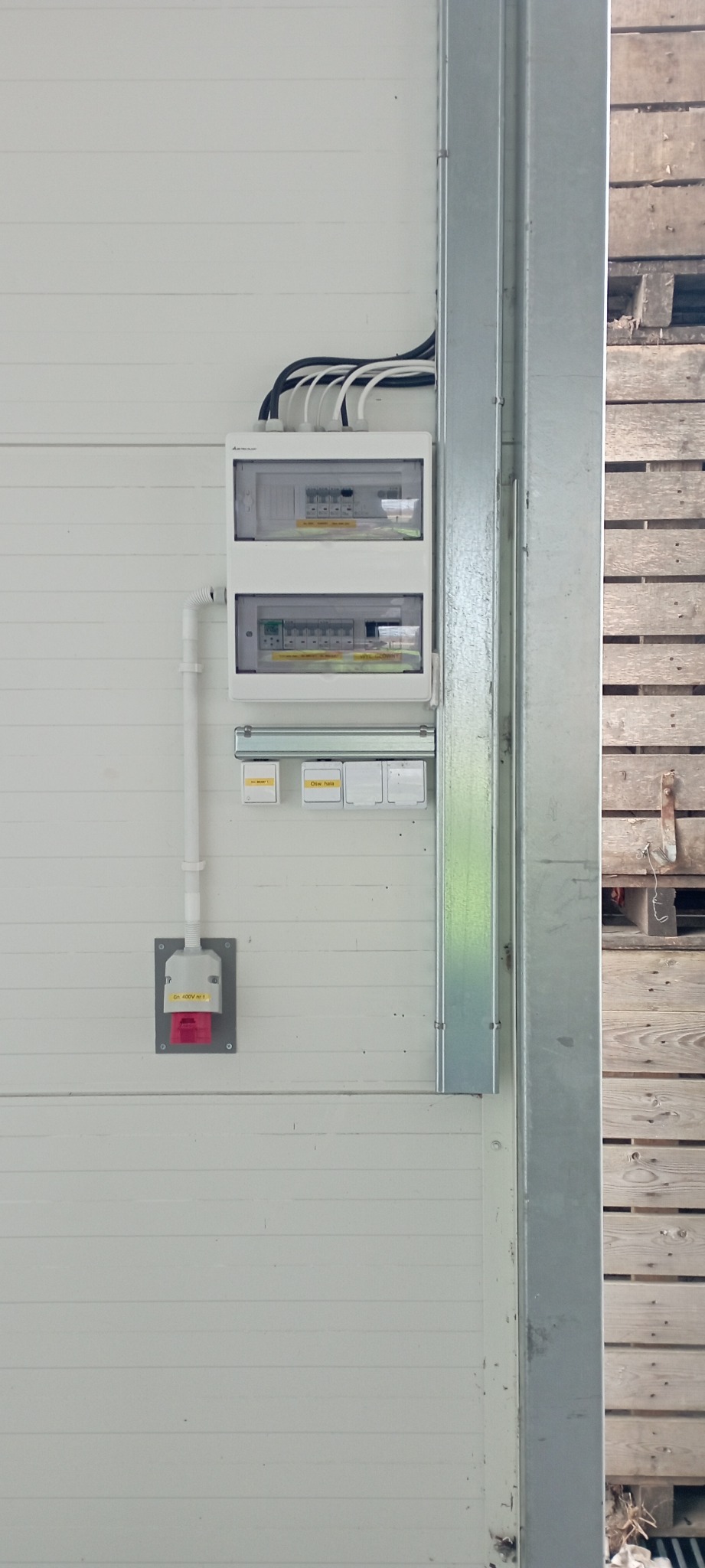 Biała skrzynka elektryczna z zabezpieczeniami i wyłącznikami, zamontowana na białej ścianie z widocznymi przewodami i gniazdem przemysłowym 400V, obok metalowej ramy i drewnianych palet.