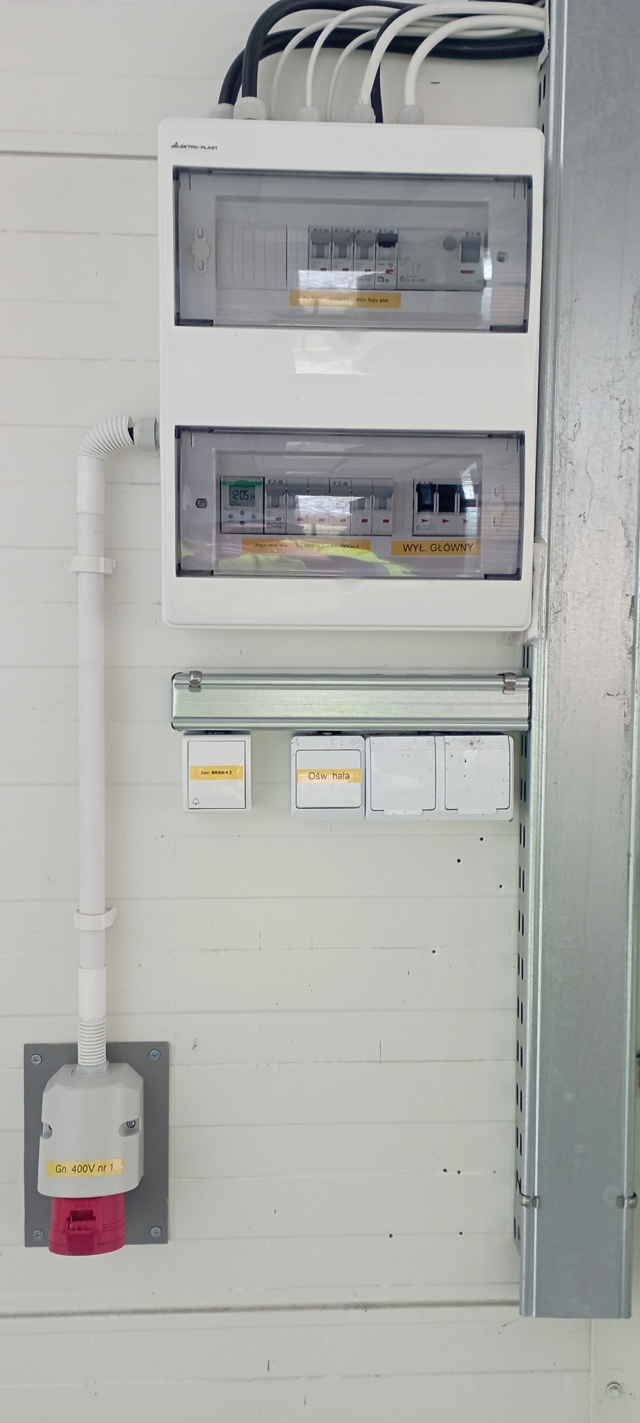 Rozdzielnica elektryczna z wyłącznikami, zabezpieczeniami i gniazdem siłowym 400V w hali, widoczne oznaczenia obwodów oświetleniowych i sterowania bramą.