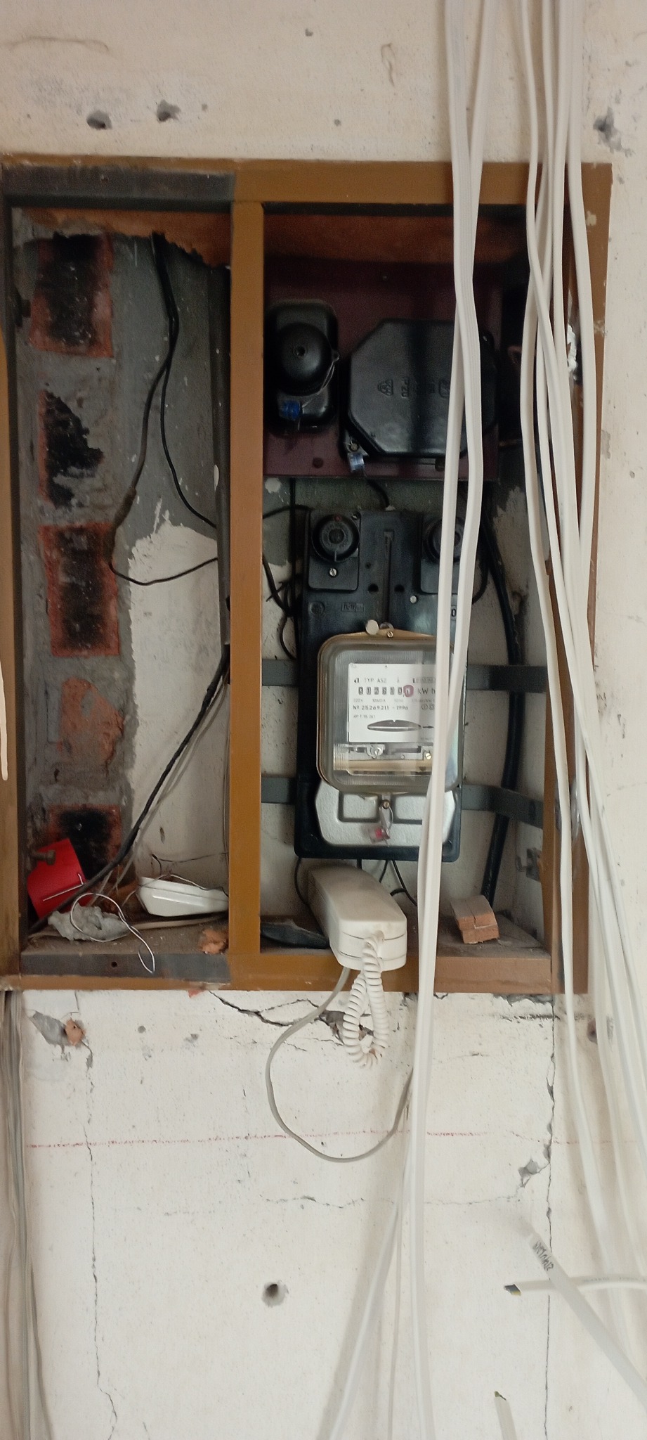 Niewykończona instalacja elektryczna: licznik w drewnianej ramie, widoczne kable i przewody, surowa ściana z cegłami i pęknięciami.