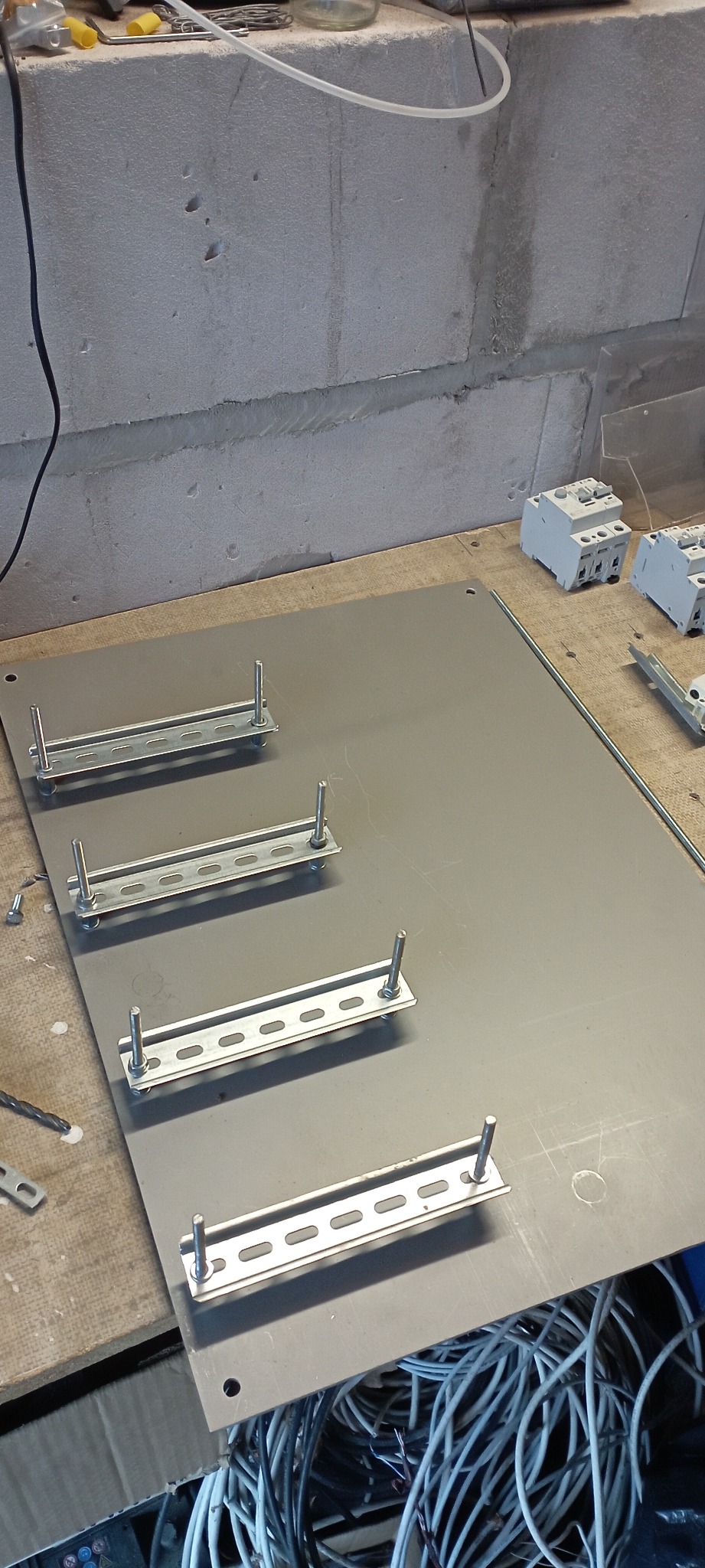 Metalowa płyta montażowa z przymocowanymi szynami DIN i śrubami, przewody elektryczne w tle.