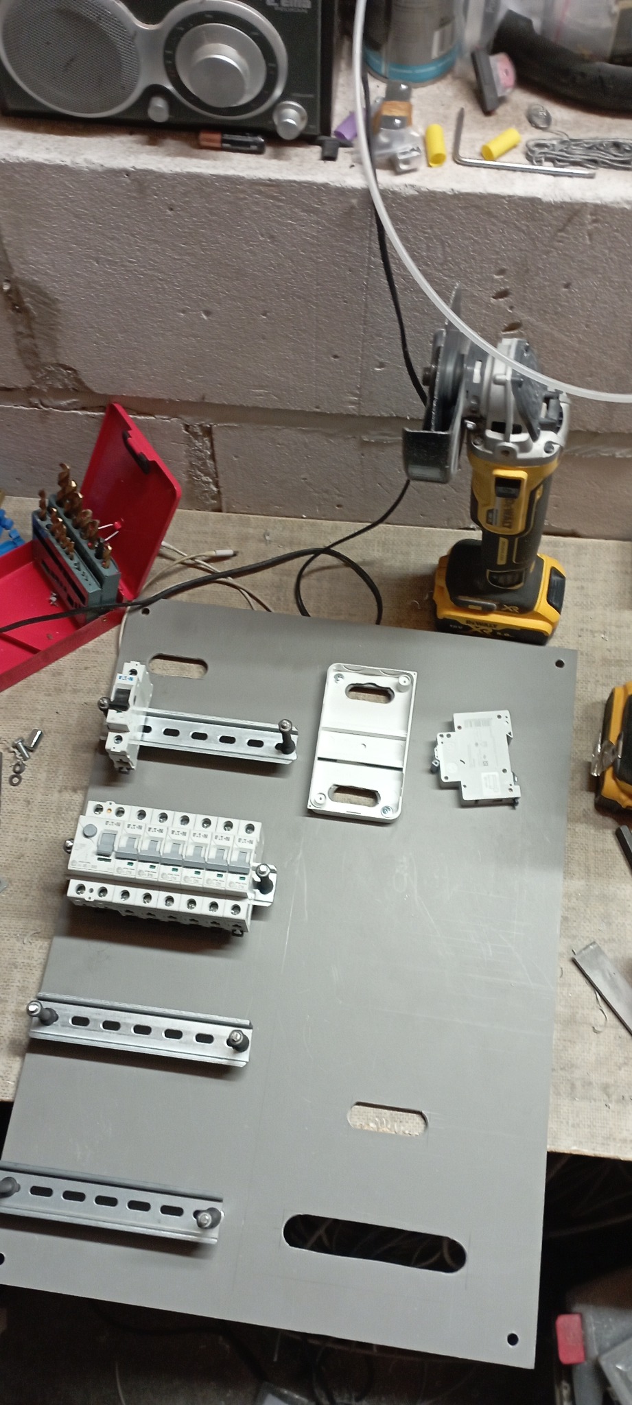 Szara płyta montażowa z zamontowanymi elementami elektrycznymi, takimi jak wyłączniki i szyny DIN, widoczne narzędzia: wiertarka akumulatorowa i otwarty pojemnik z wiertłami, na warsztatowym stole...