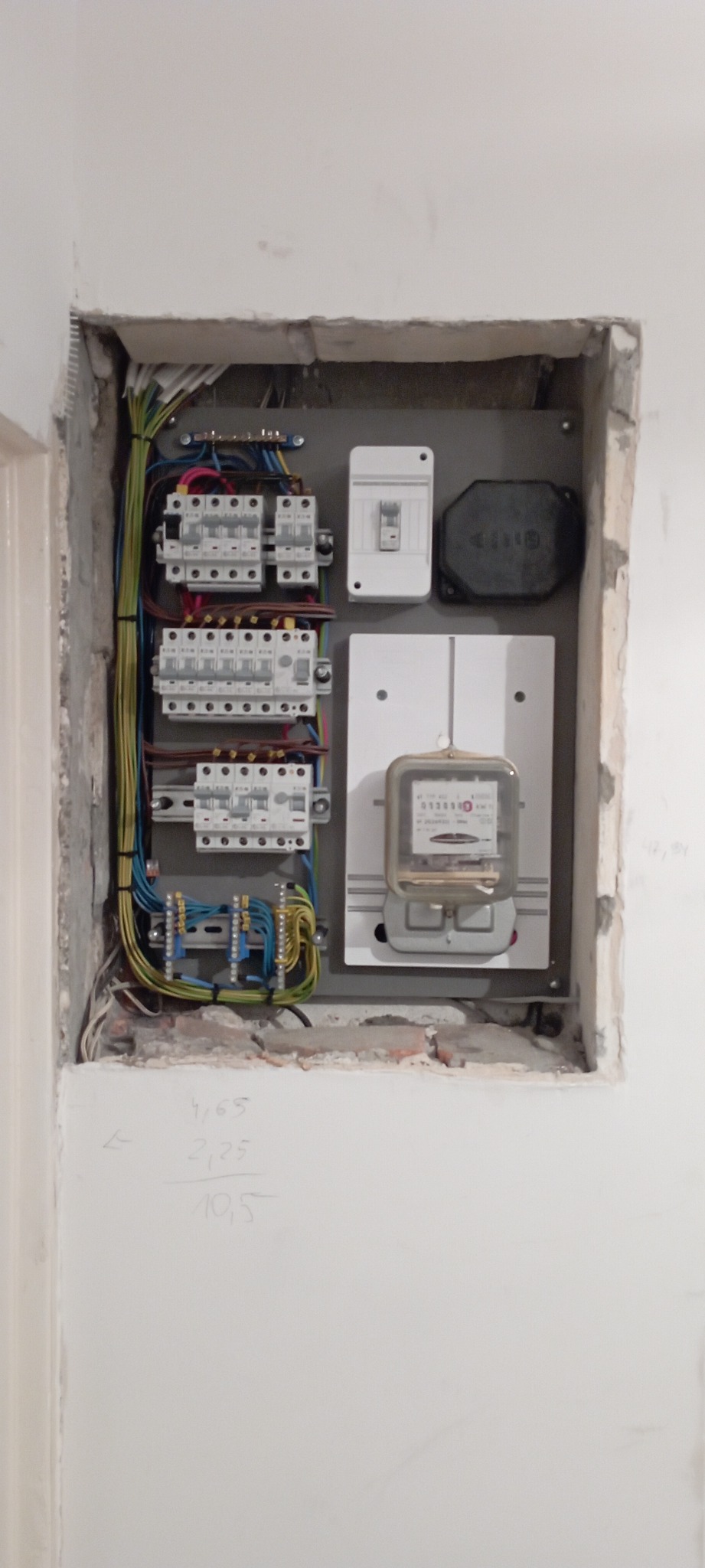 Wnęka w ścianie z zamontowaną skrzynką elektryczną, widoczne kable, bezpieczniki i licznik energii elektrycznej. Ściana wokół wnęki z widocznymi śladami prac murarskich i zapisanymi ołówkiem liczbami.
