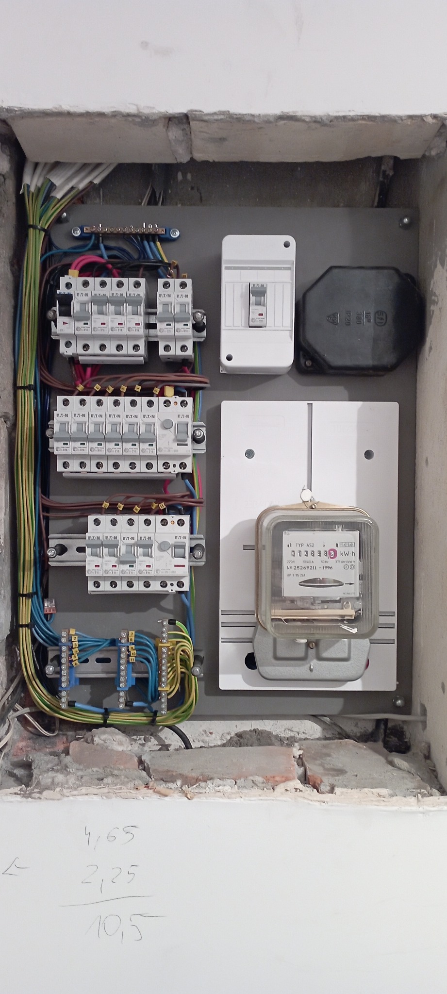 Skrzynka elektryczna z bezpiecznikami, wyłącznikiem i licznikiem energii elektrycznej, zamontowana w surowym otworze w ścianie, widoczne kable i instalacja. Na ścianie poniżej otworu widoczne...