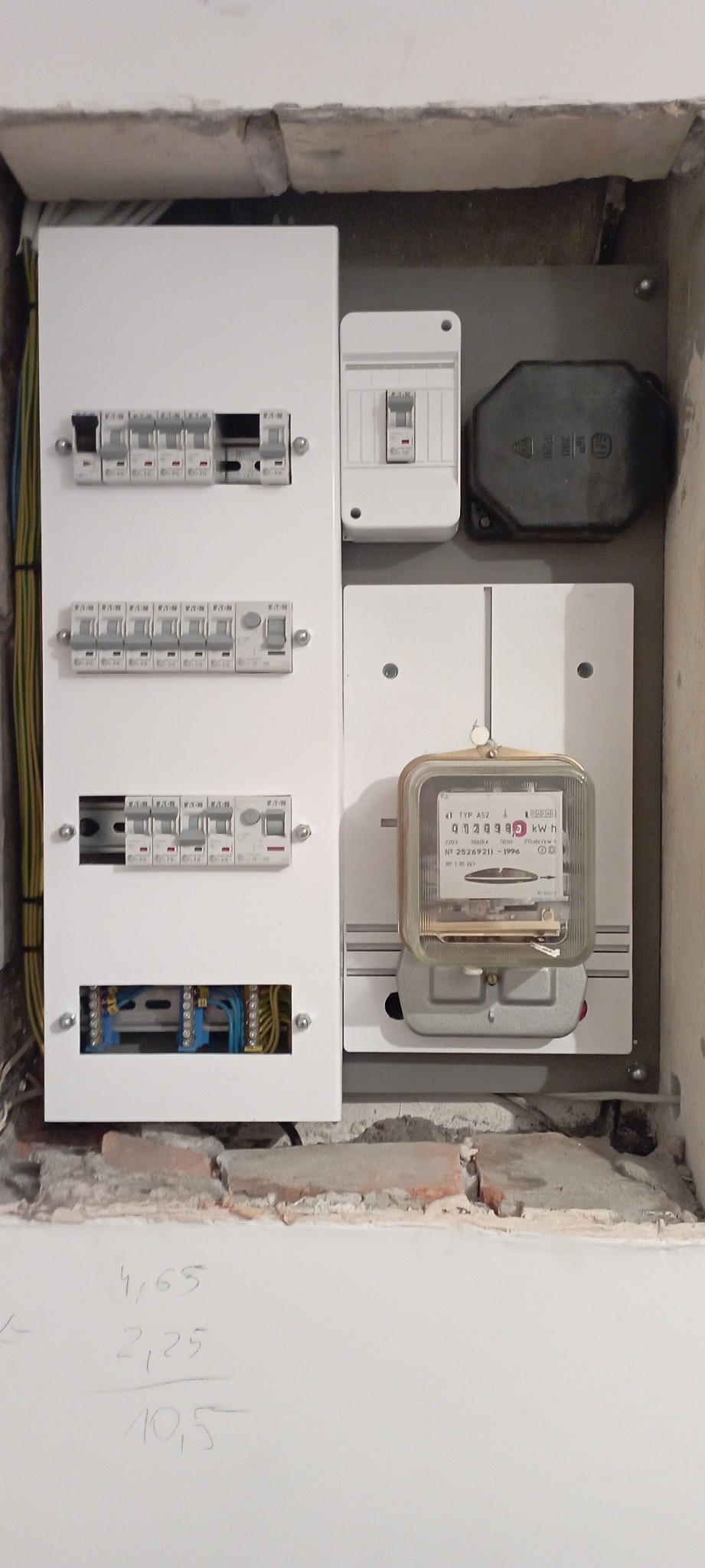 Tablica rozdzielcza z bezpiecznikami i licznikiem energii elektrycznej zamontowana we wnęce w ścianie, widoczne przewody i instalacja elektryczna.