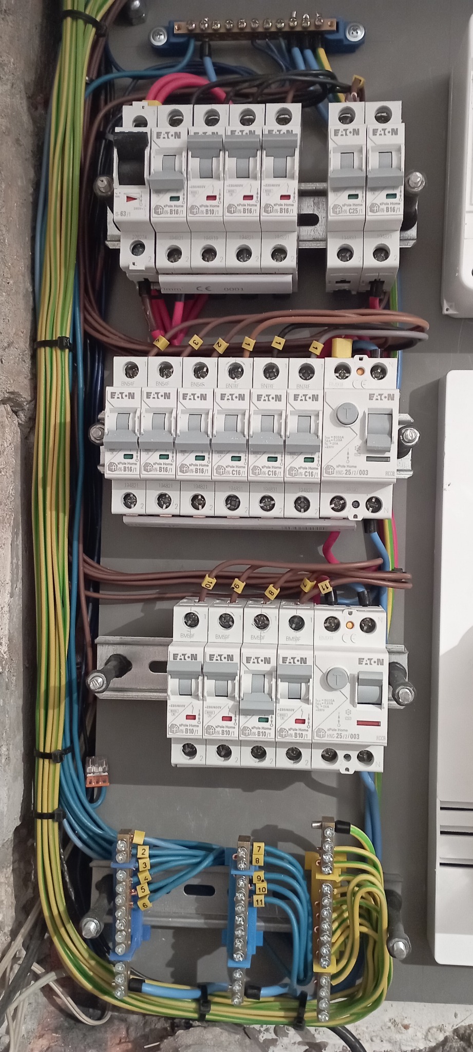 Rozdzielnica elektryczna z zamontowanymi bezpiecznikami marki Eaton, okablowaniem oznaczonym numerami i szynami zbiorczymi, widok z bliska na instalację w szarej obudowie.