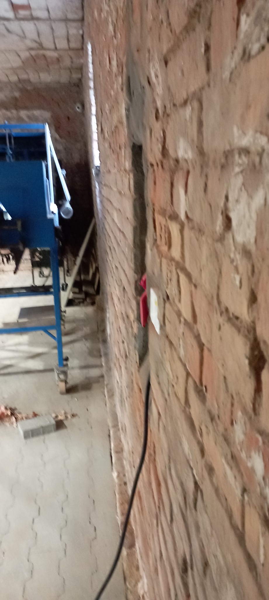 Wnętrze budynku gospodarczego w trakcie rozbudowy instalacji elektrycznej, widoczny kabel elektryczny prowadzony w wyżłobieniu w ceglanej ścianie, z fragmentem niebieskiego urządzenia w tle.