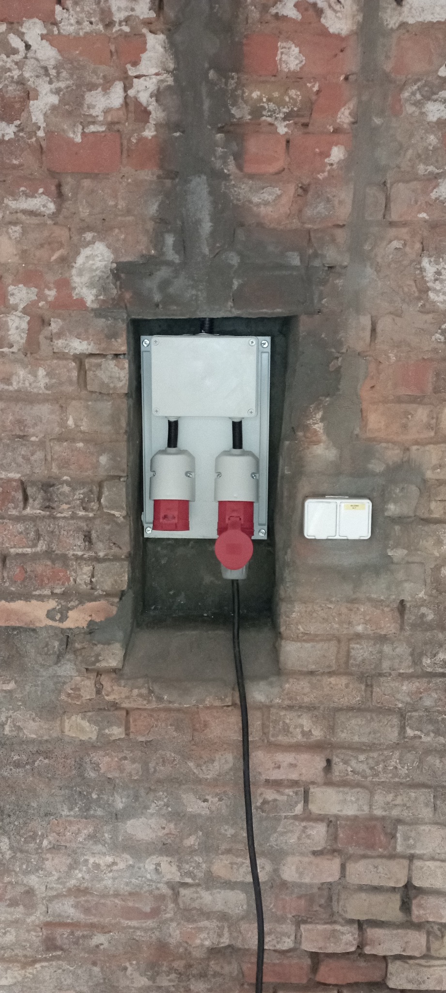 Gniazda siłowe (czerwone wtyczki) zamontowane w puszce na ceglanej ścianie z widocznymi śladami zaprawy, obok gniazdko podwójne.