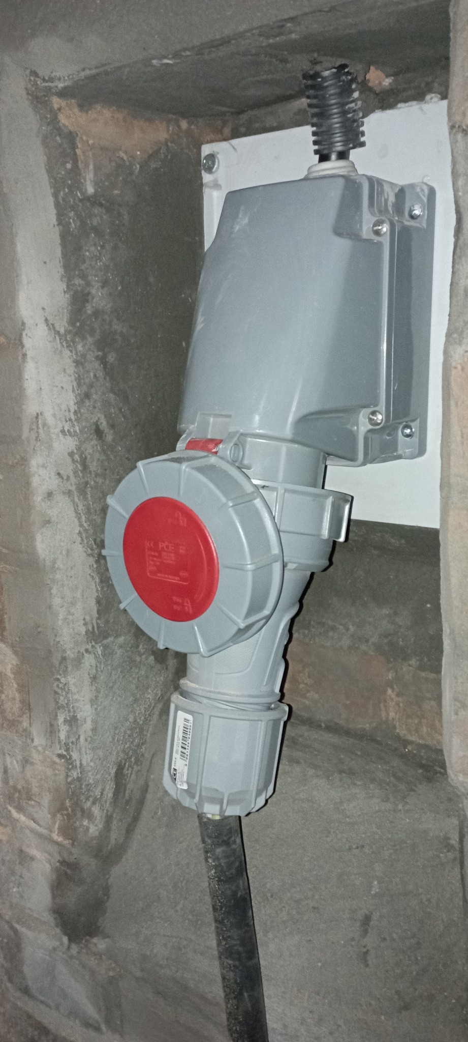 Szare gniazdo siłowe z czerwoną pokrywą zamontowane na ścianie, widoczny czarny kabel w szorstkim, betonowym otoczeniu.