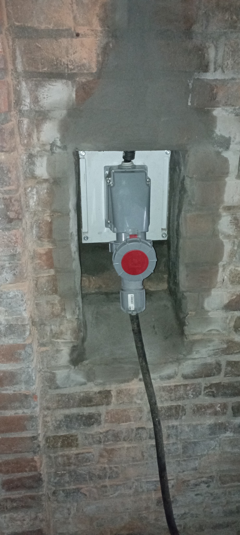 Szare gniazdo przemysłowe z czerwonym przyciskiem zamontowane na białej skrzynce na tle ceglanej ściany z widocznymi śladami zaprawy.