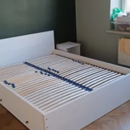 Rama łóżka z drewna bielonego z listwami i dwoma szafkami nocnymi ustawiona w pokoju z parkietem i zielonymi ścianami, widok na okno z parapetem zagraconym przedmiotami.